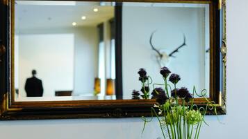 Un espejo con un marco decorativo que refleja el interior de una habitación, un jarrón con flores y una persona en el reflejo.
