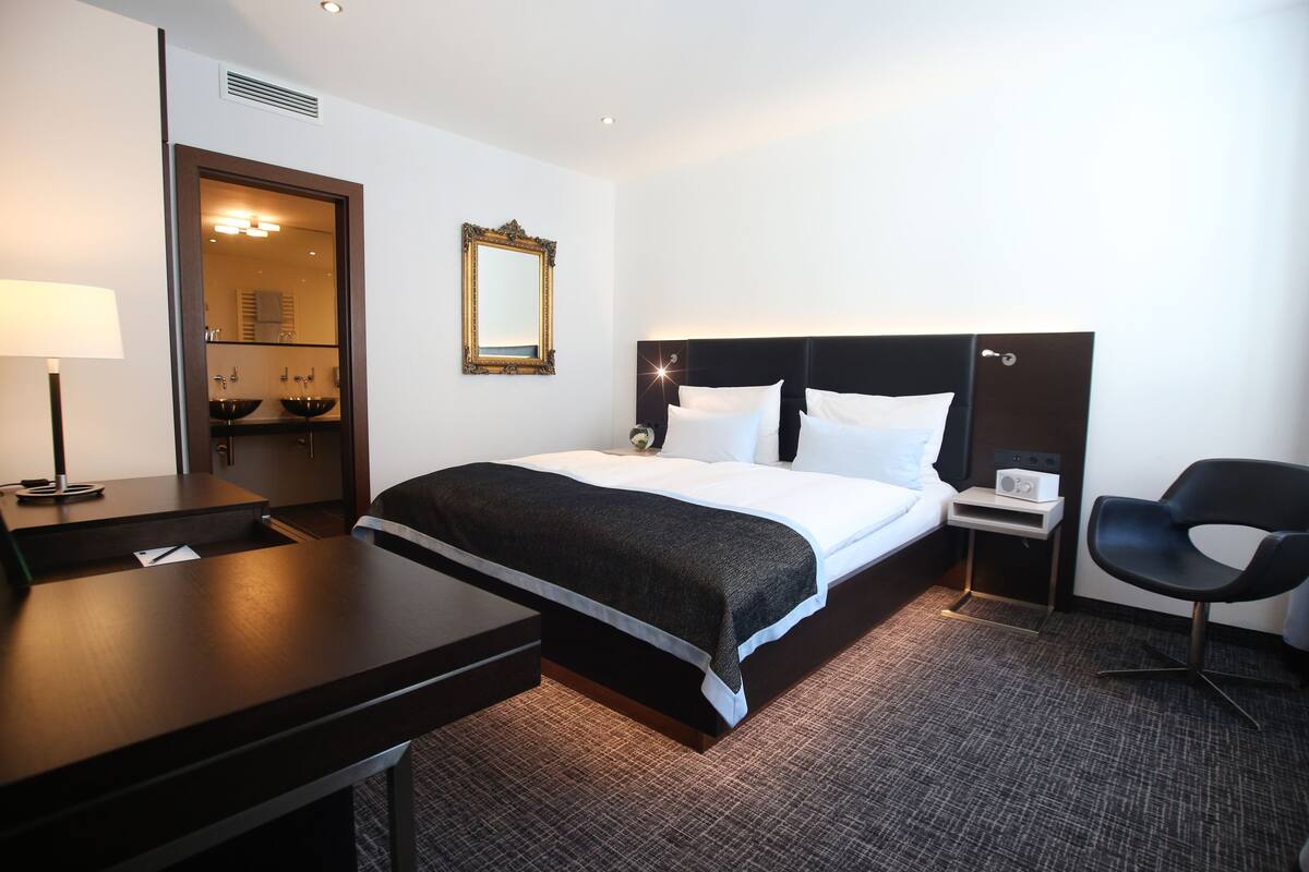 A hotel room with a bed, bedside table, chair, and a mirror.