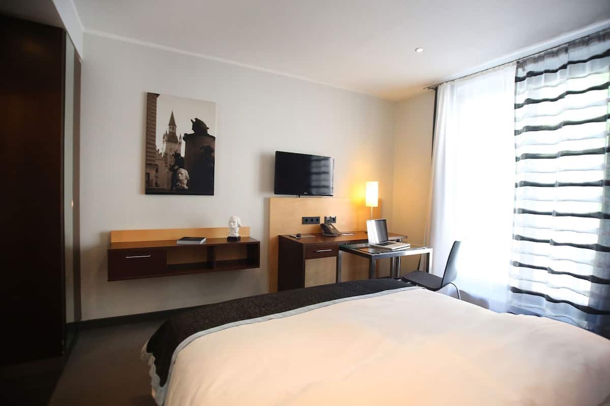 A hotel room with a bed, a desk with a chair, a television, and a window with blinds.