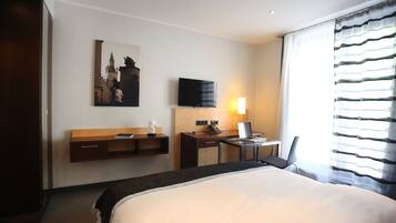 A hotel room with a large bed, a desk with a computer, a flat-screen TV, a small table with a chair, and a wall-mounted shelf with a statue.