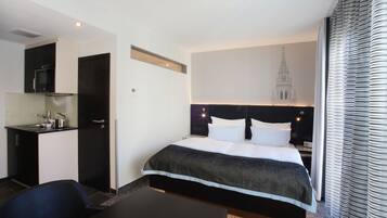 A hotel room with a bed, a desk, a kitchenette, and a view of a building.