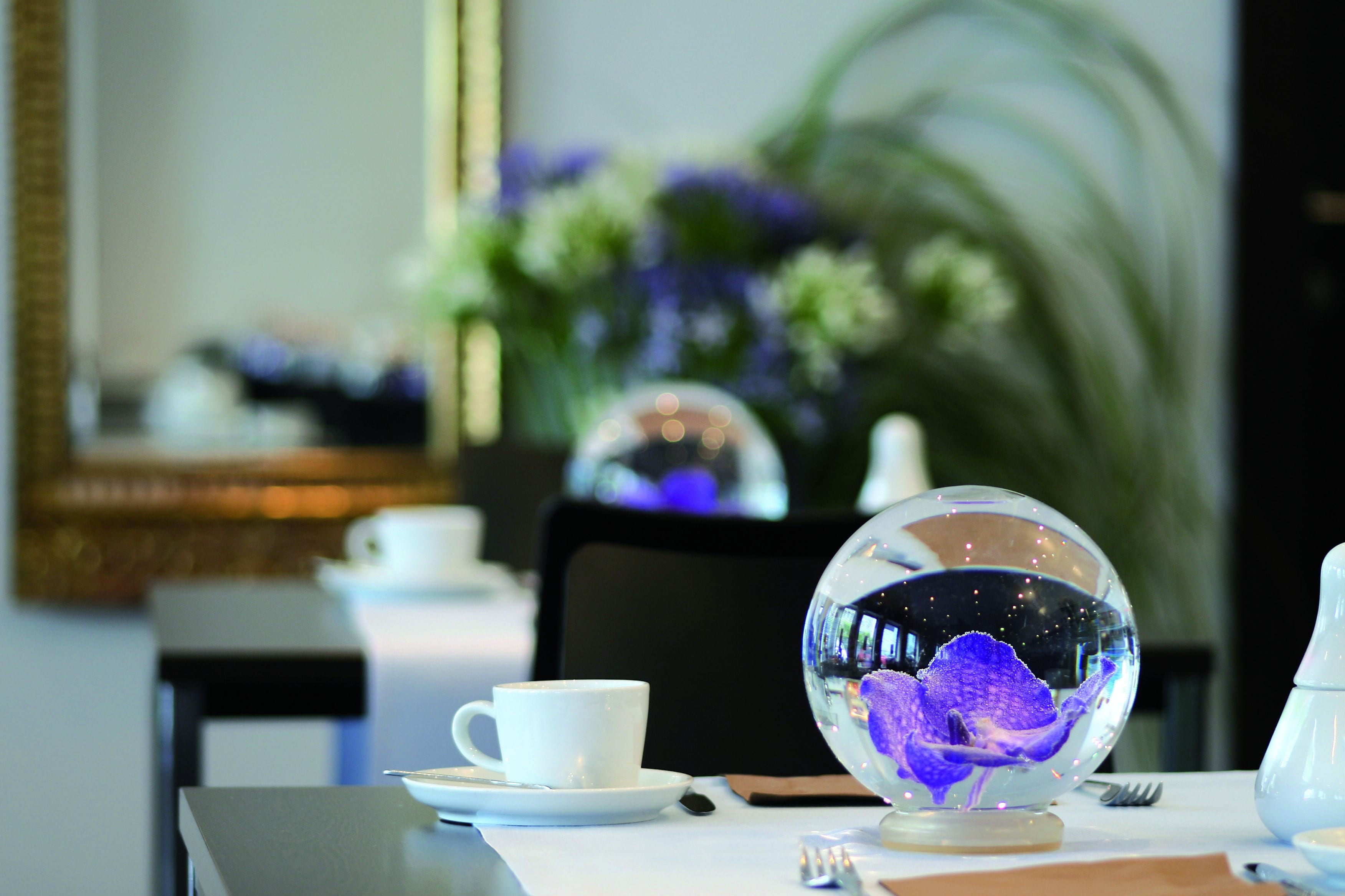 Una mesa de comedor con una taza blanca, un plato bajo y un cuenco de vidrio transparente que contiene una flor morada.