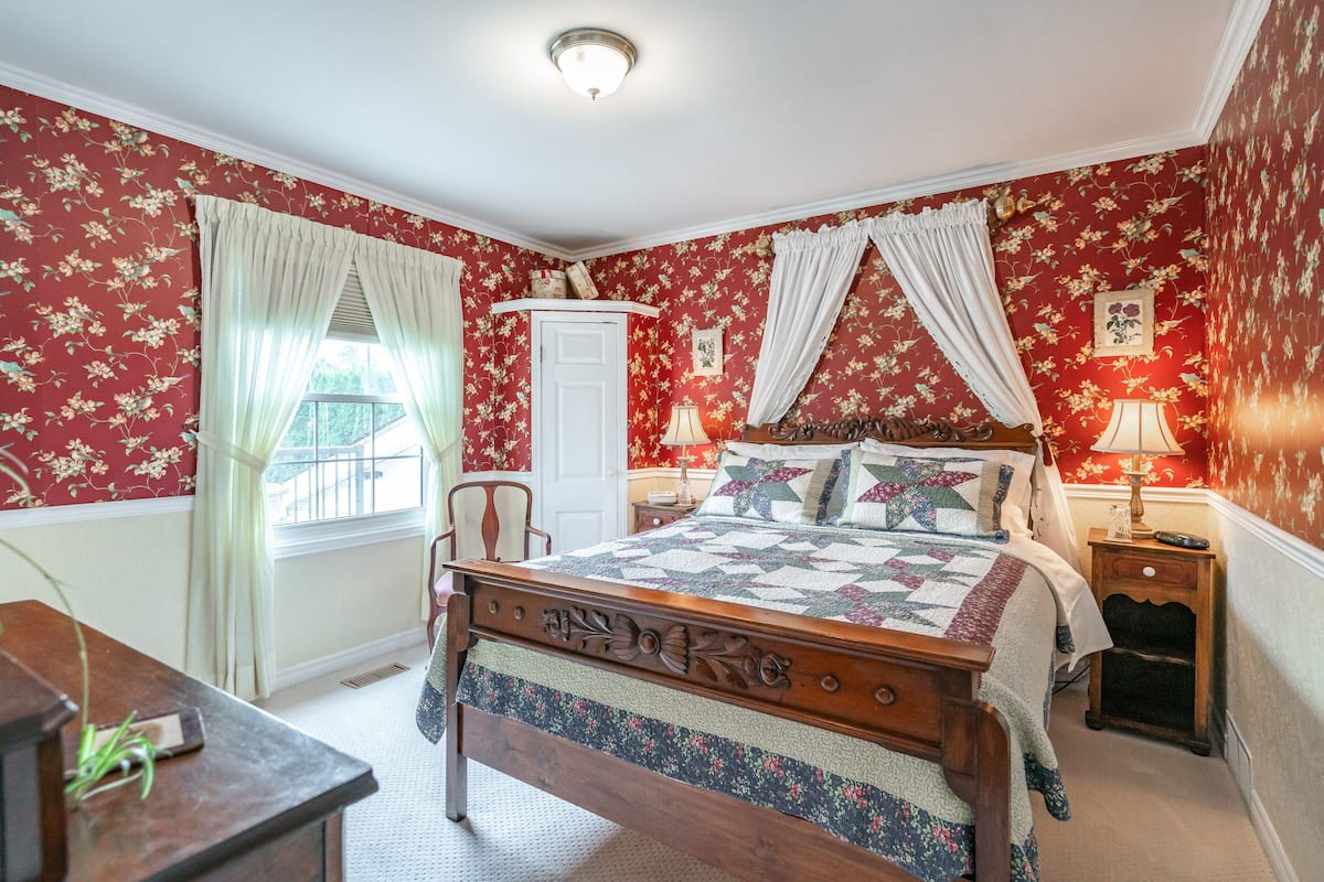 A bedroom with a floral wallpaper, a wooden bed with a canopy, a nightstand with a lamp, and a chair.