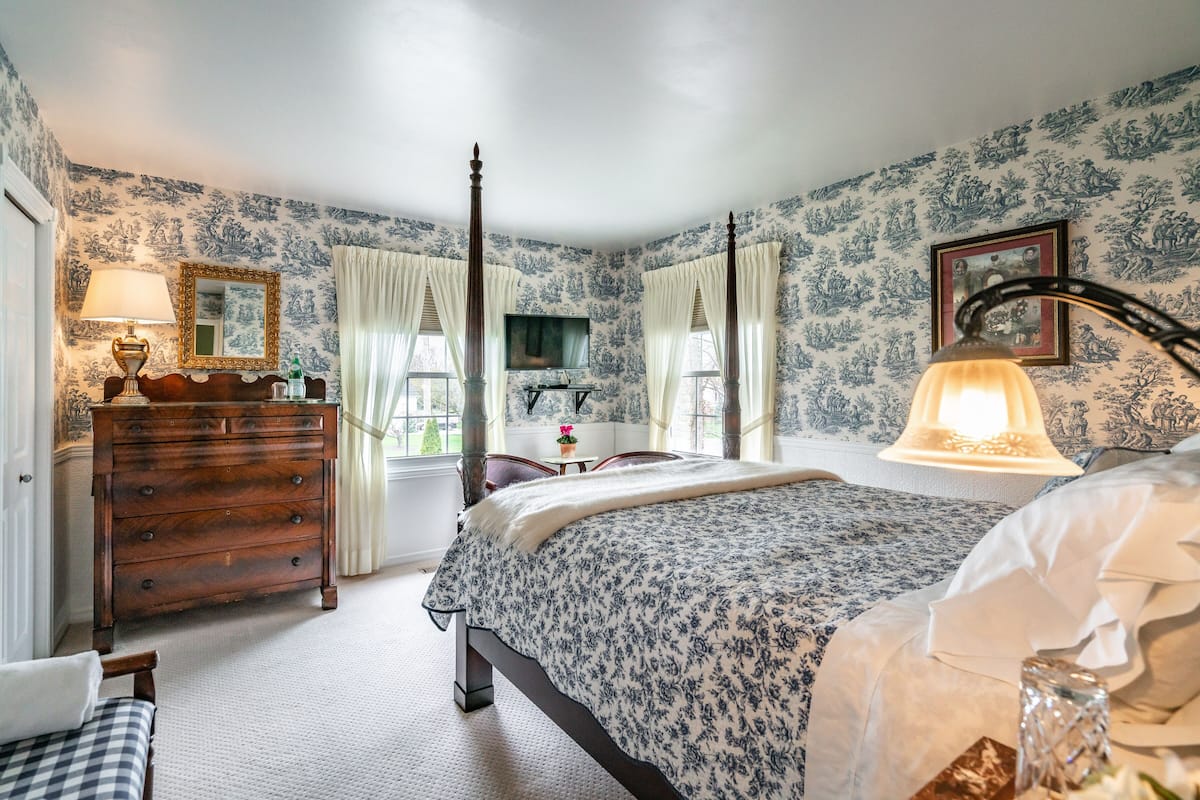 A bedroom with a four-poster bed, a wooden dresser, a vanity with a mirror, and a window with curtains.
