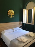 Un letto rifatto con lenzuola bianche e asciugamani piegati, una testata in legno, una decorazione a forma di albero appesa al muro e una finestra con persiane.