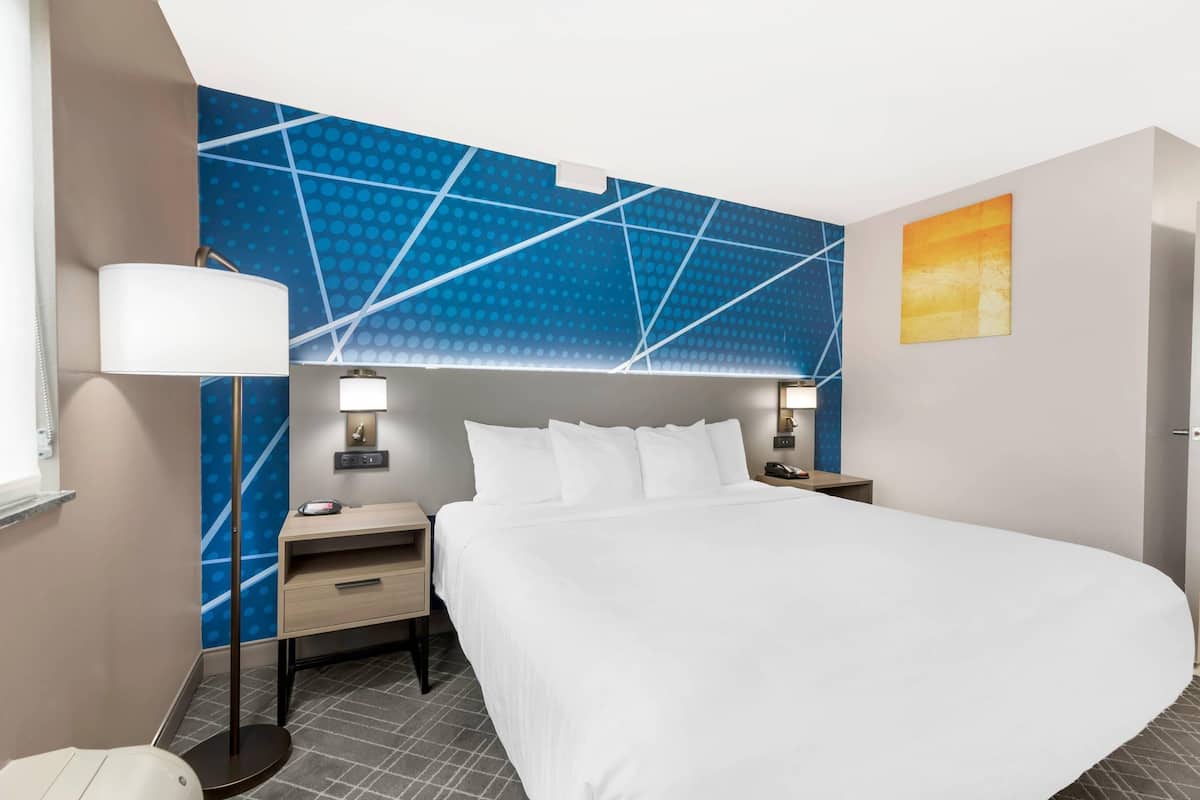 Une chambre d’hôtel avec un grand lit, un bureau, une chaise, une lampe, une table de chevet et un mur orné d’un motif géométrique.
