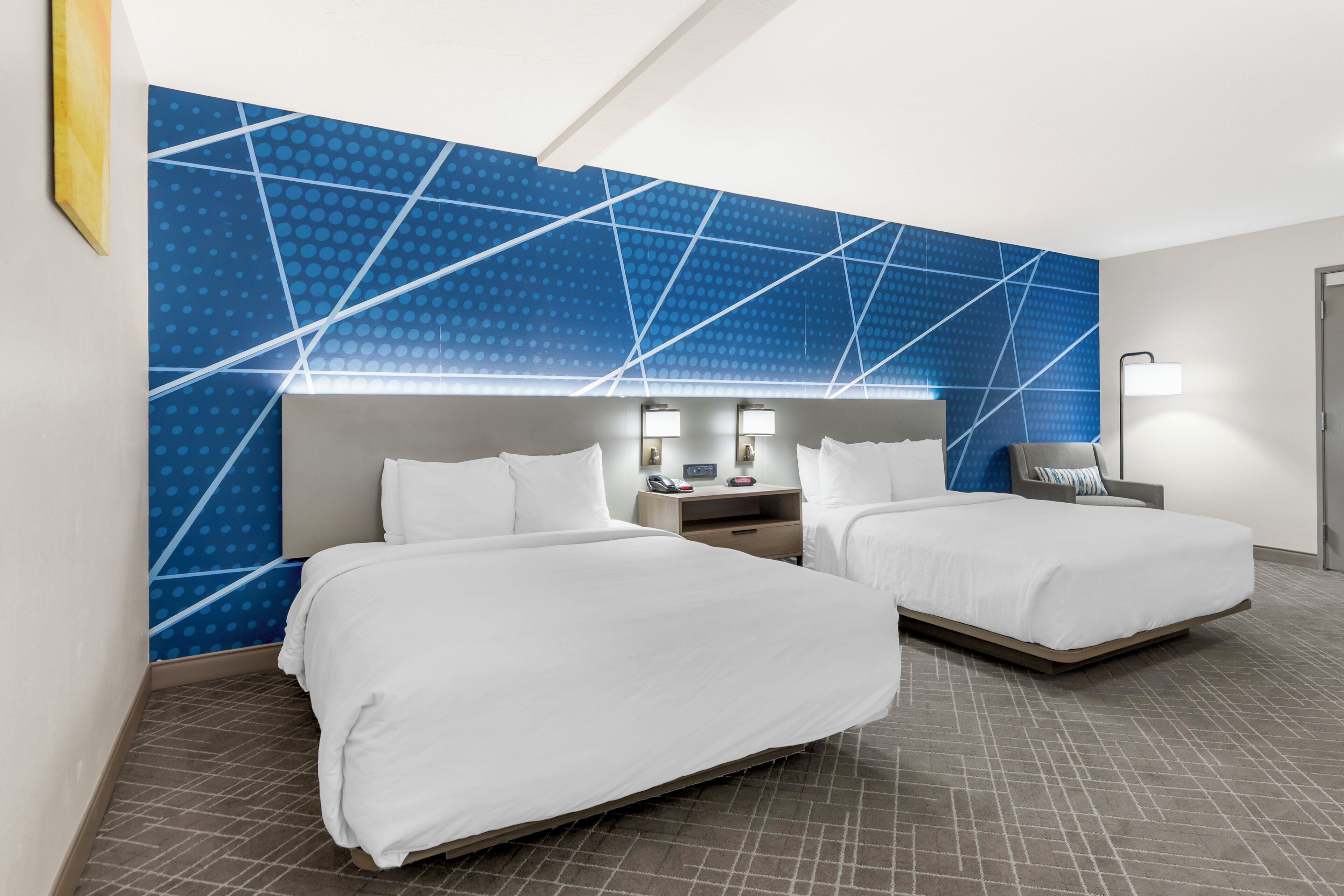 Et hotelværelse med to senge, et blåt geometrisk vægdesign og en moderne lampe.