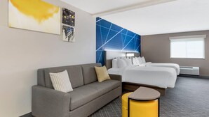 Une chambre d’hôtel moderne avec un canapé gris, un lit avec du linge de lit blanc, un petit pouf jaune et des œuvres d’art accrochées aux murs.