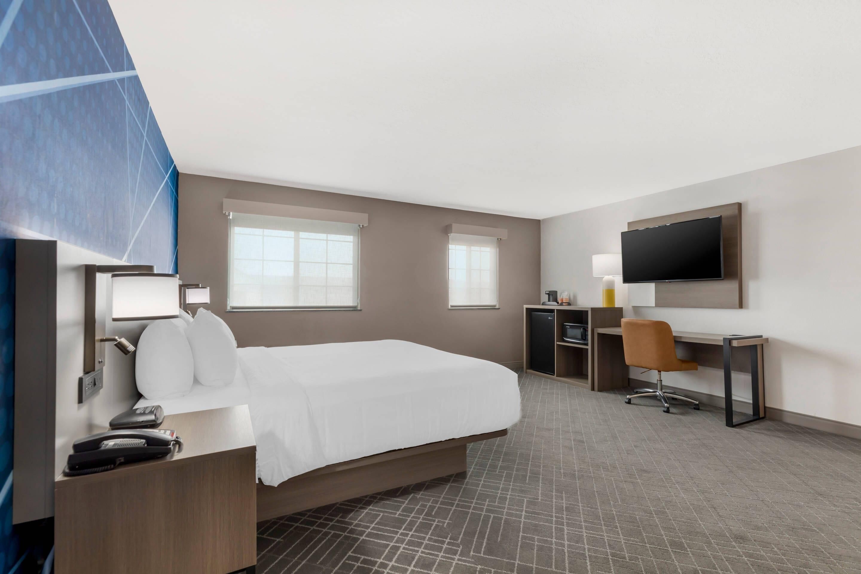 A hotel room with a bed, desk, chair, TV, and a small kitchenette.