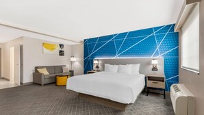Une chambre d’hôtel moderne avec un grand lit, un canapé gris, une petite table et un mur orné d’un motif géométrique.