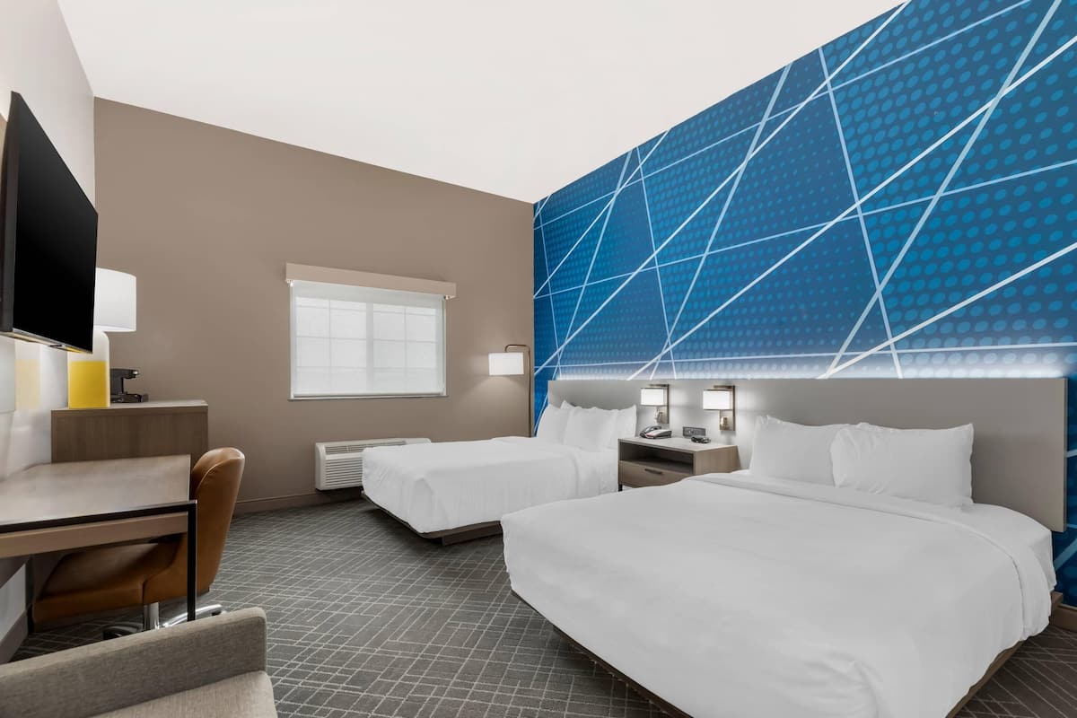 Une chambre d’hôtel avec deux lits, un bureau, une chaise, une télévision et un grand mur orné d’un motif géométrique.