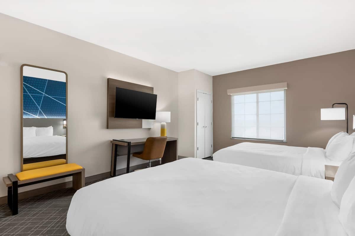 Une chambre d’hôtel avec un grand lit, un lit plus petit, un bureau avec une chaise, une télévision et une fenêtre avec des stores.