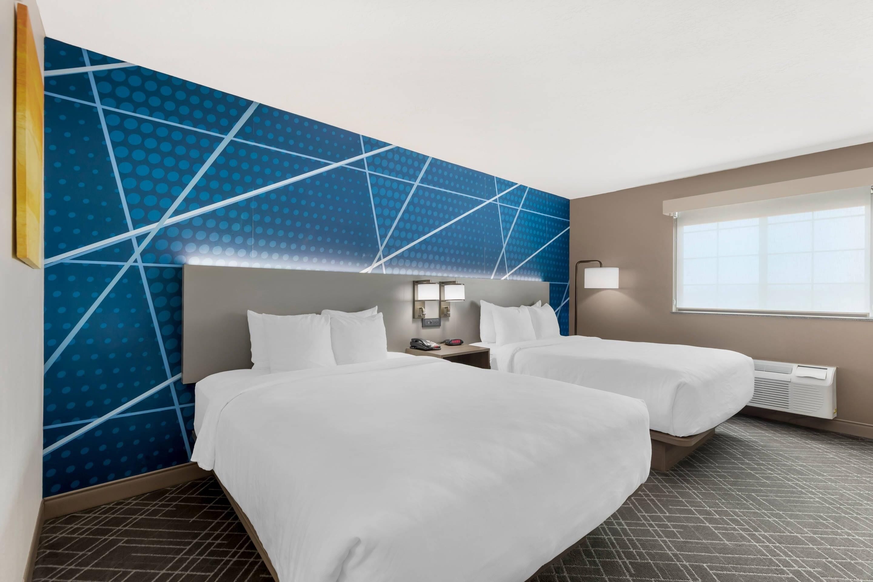 A hotel room with two beds, a blue patterned wall, a window with blinds, and a wall-mounted heater.