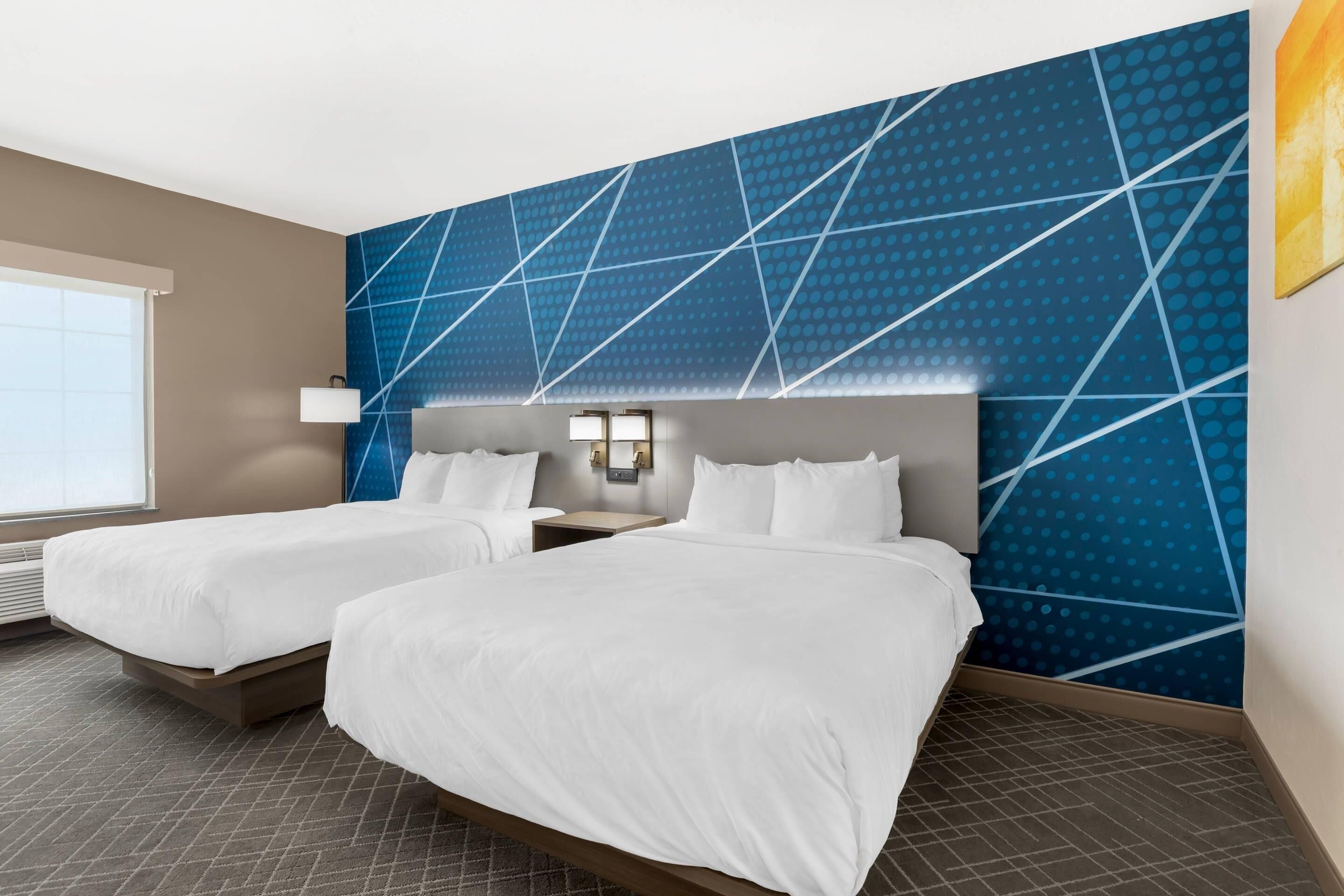 A hotel room with two beds, a large window, and a blue geometric wall design.