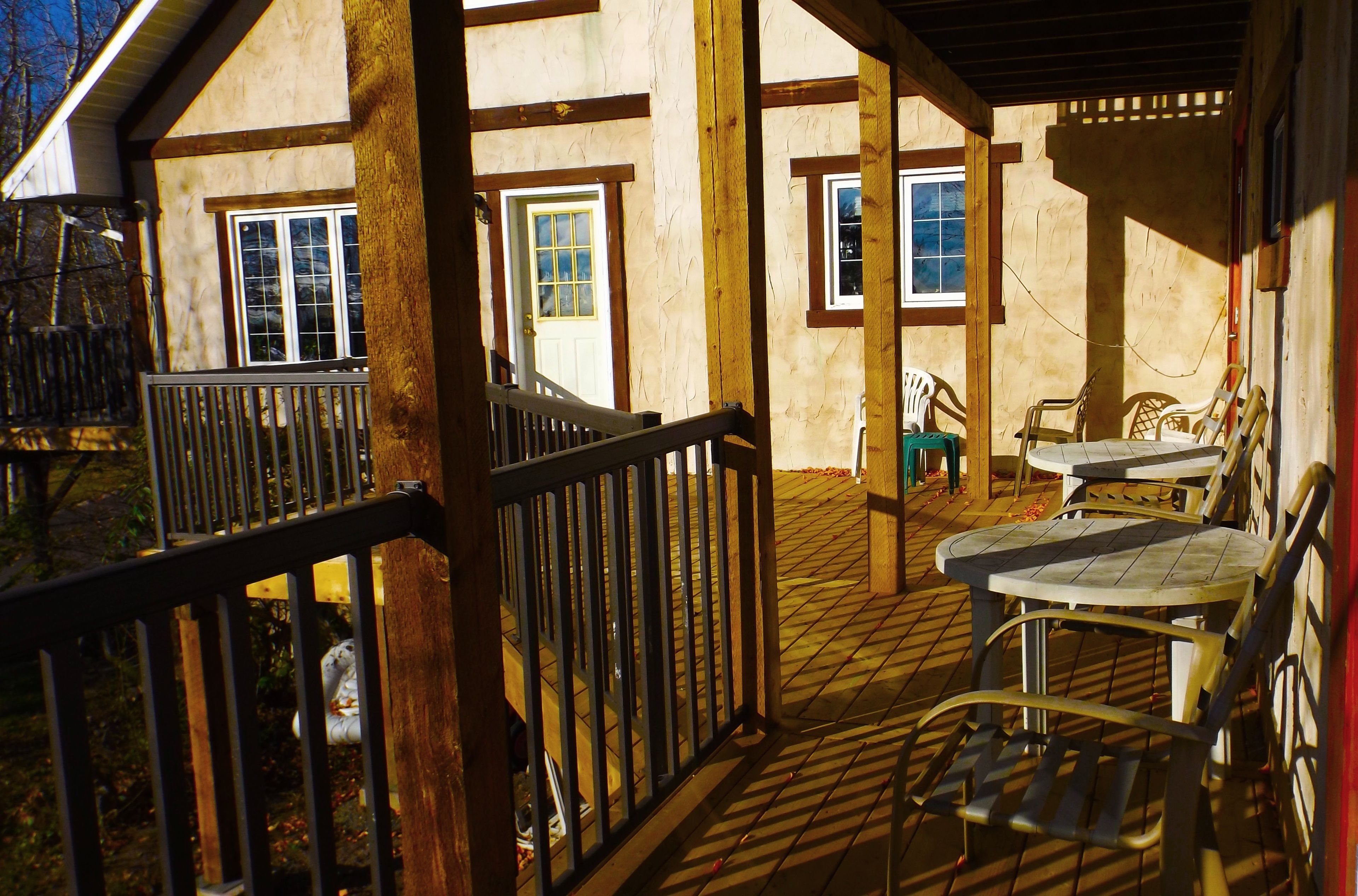 Un espacio al aire libre techado con una terraza de madera, una mesa con sillas y vistas a una casa con ventanas y una puerta.