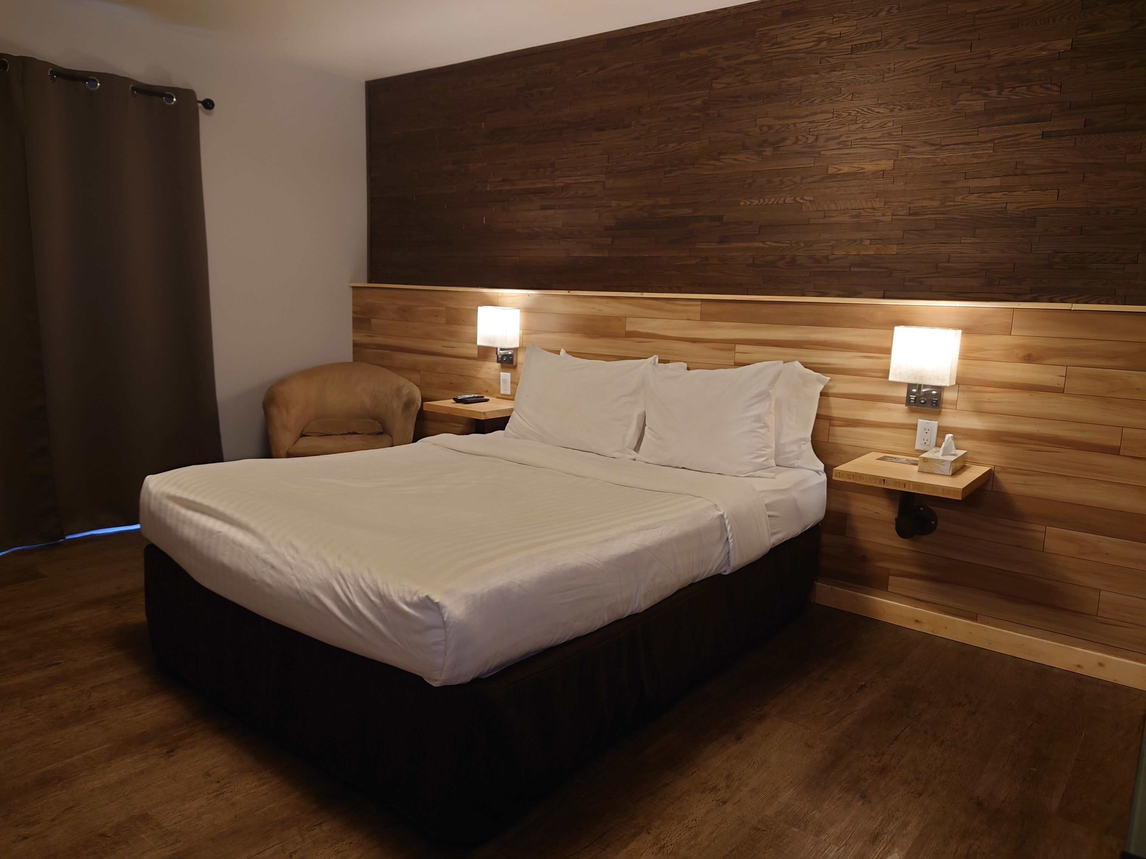 A hotel room with a wooden headboard, a bed with white linens, two bedside lamps, a chair, and a nightstand with a tissue box.