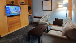 A cozy living room with a wooden ceiling, a flat-screen TV, a sofa, a patterned armchair, and a small table. - Timberland Inn (Kenora)
