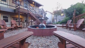 A cozy outdoor seating area with Adirondack chairs, a fire pit, and a staircase leading to a building with multiple windows and a balcony. - Timberland Inn (Kenora)