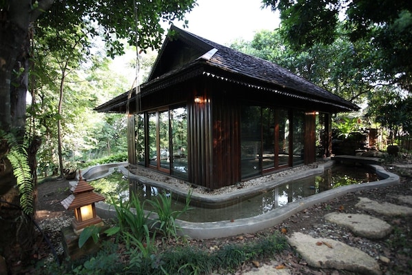 A traditional wooden structure with a curved pond, surrounded by lush greenery and stone pathways.