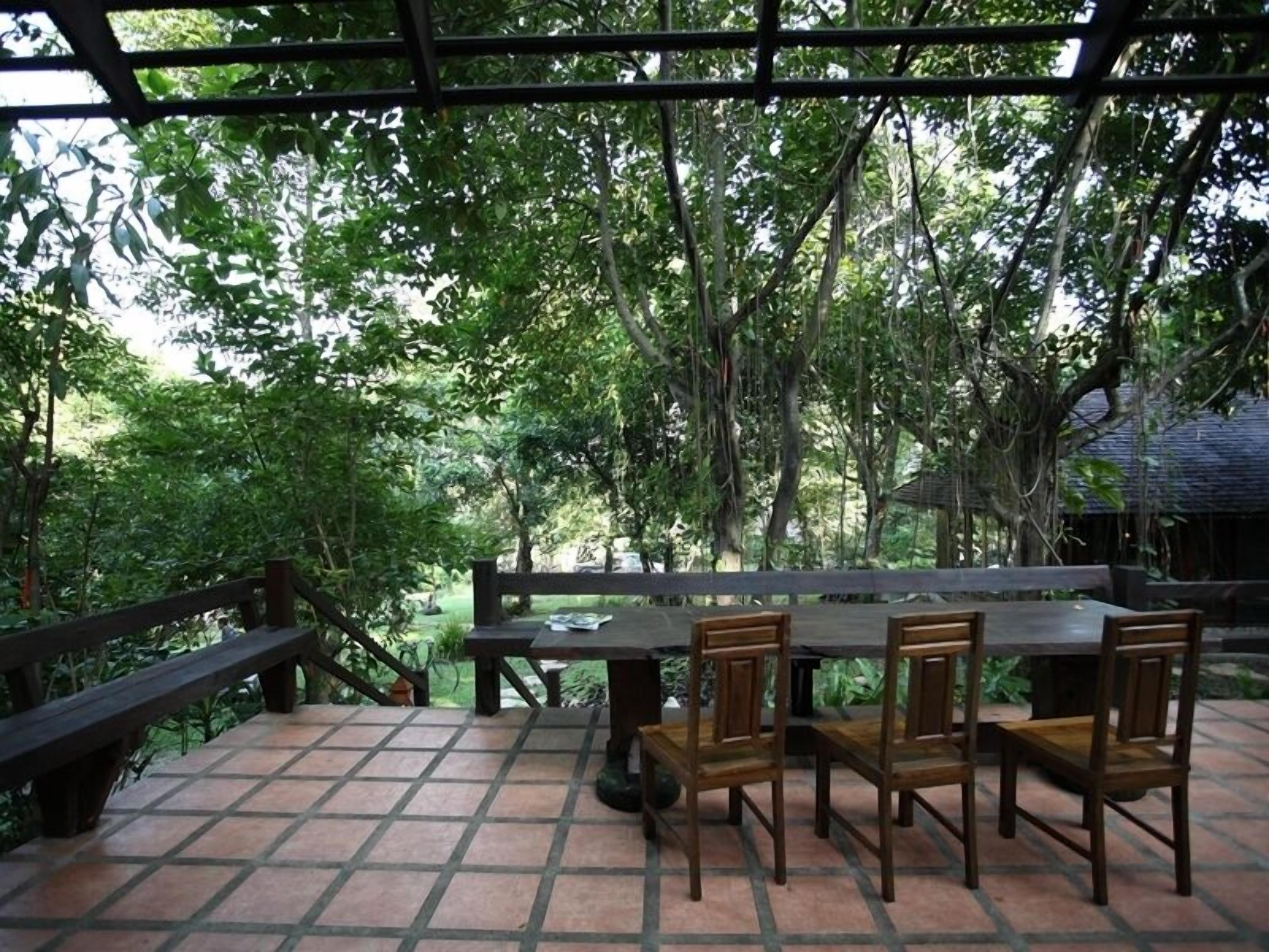 A covered outdoor dining area with a wooden table and chairs, surrounded by lush greenery and trees.