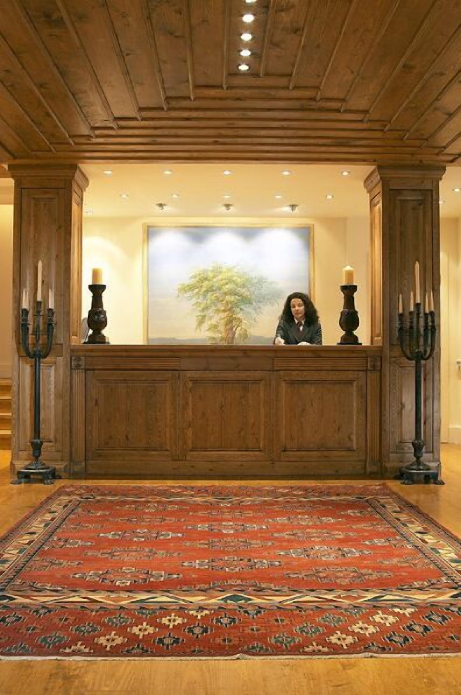 Un mostrador de recepción con detalles de madera, un cuadro grande enmarco de un árbol, y una alfombra roja estampada en el suelo.