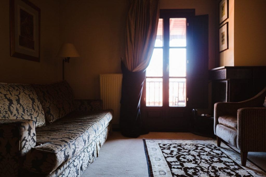 Una habitación acogedora con un sofá estampado, una mesita y una silla. Un ventanal con cortinas deja entrar luz natural.