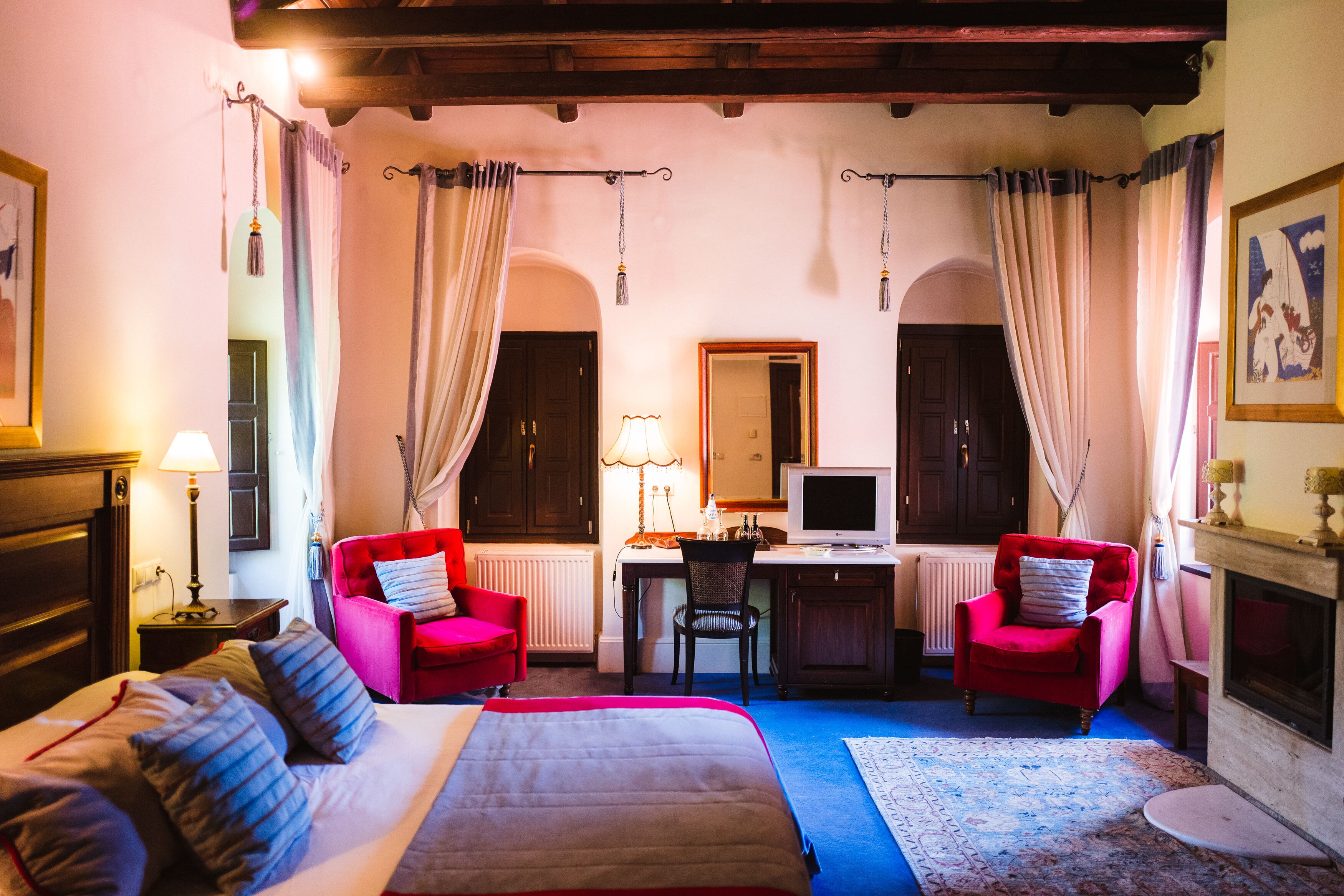 Habitación de hotel con cama, dos sillones rojos, escritorio con ordenador, chimenea y un espejo grande.