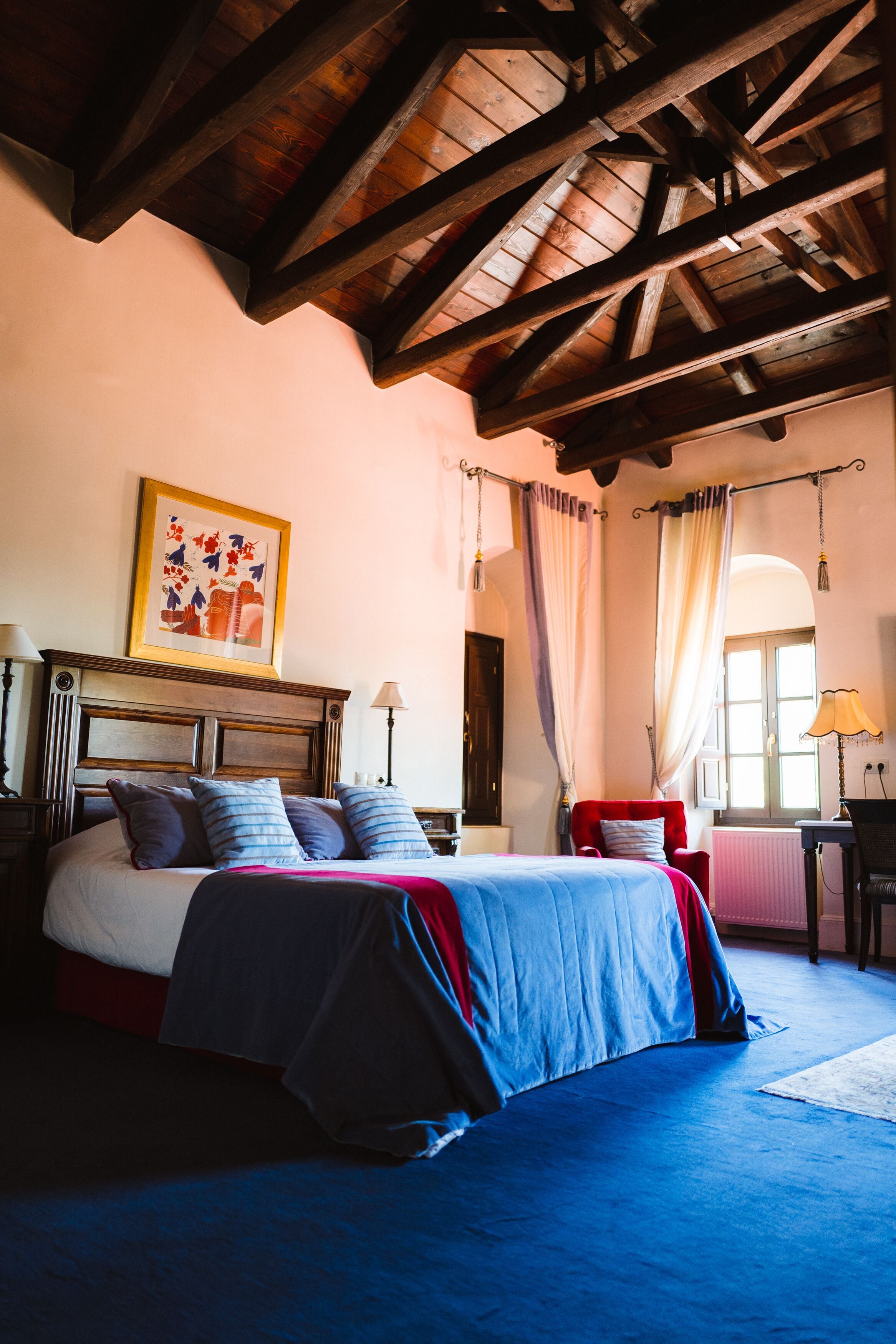 A bedroom with a wooden ceiling, a bed with a blue and red bedspread, a red chair, and a window with curtains.