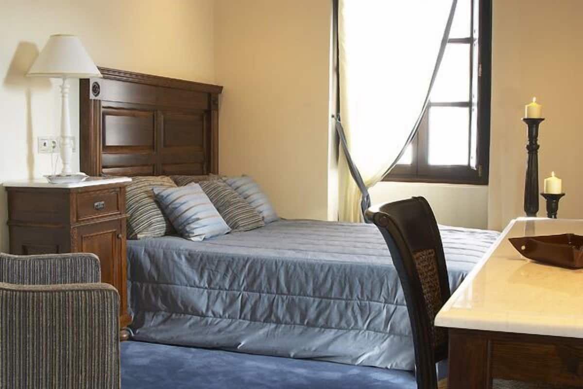 A bedroom with a bed, bedside table, lamp, chair, and window.