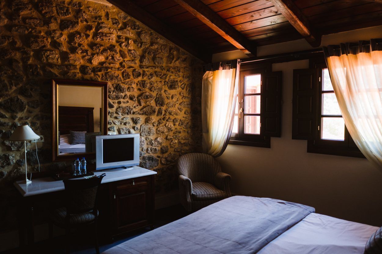 Une pièce avec un mur en pierre, un lit, un bureau avec un ordinateur, une chaise et une fenêtre avec des rideaux.