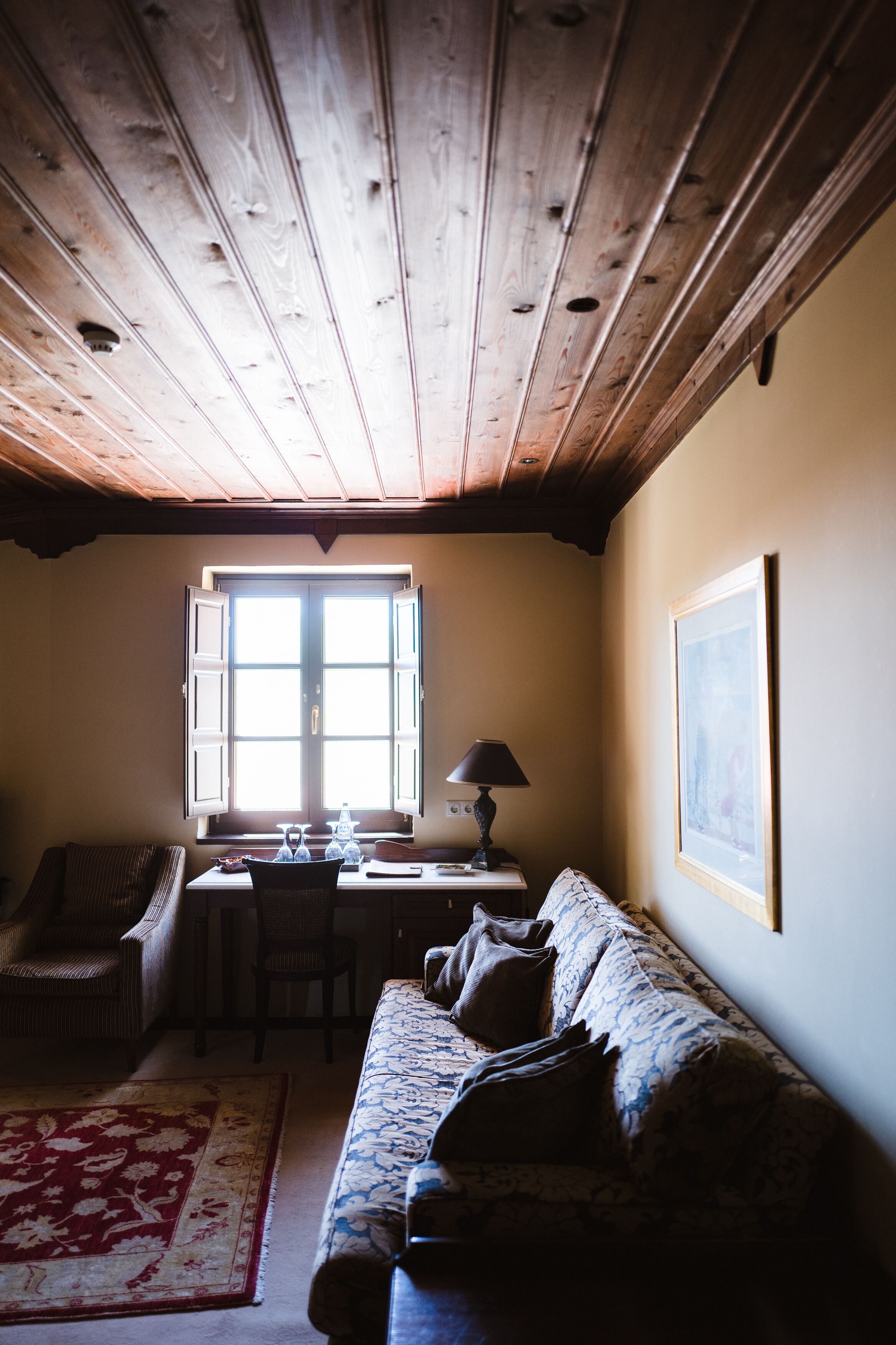 Una camera accogliente con soffitto in legno, una finestra con persiane aperte, un divano, una scrivania con una lampada e un quadro incorniciato appeso al muro.