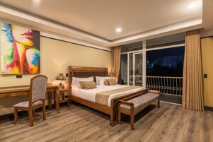 A hotel room with a large bed, a desk with a chair, a nightstand, and a view of the city at night.
