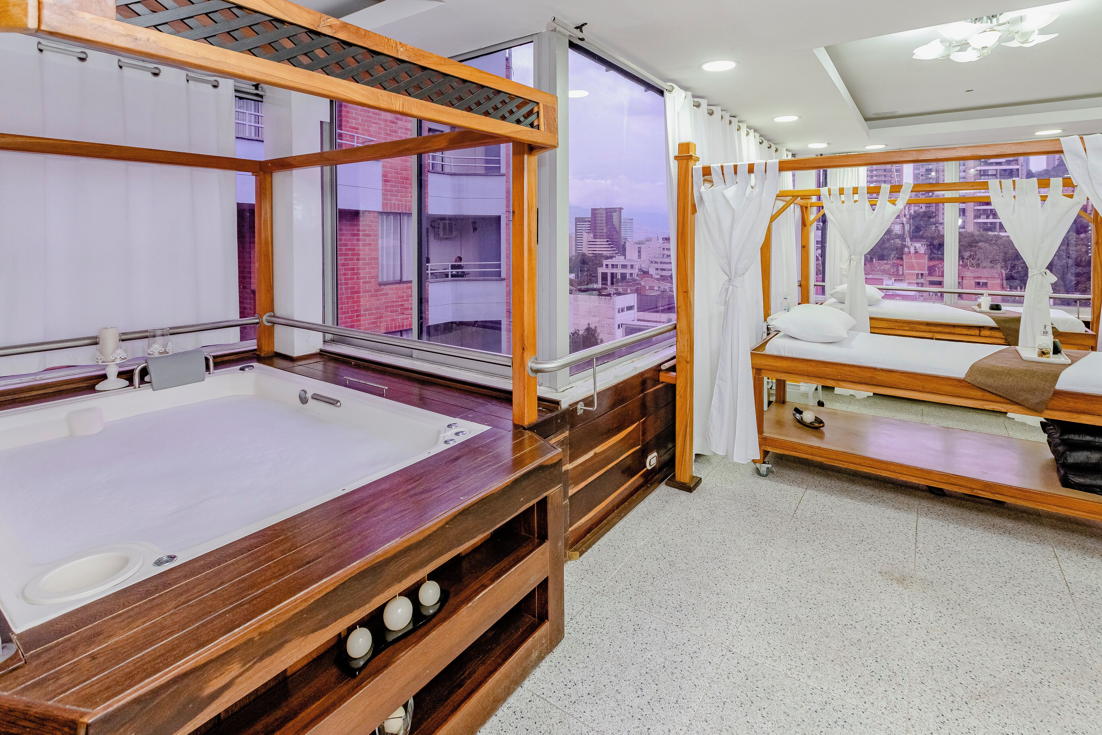 A bedroom with a wooden bed, a large window with curtains, a wooden bedside table, and a view of buildings outside.
