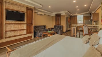 A hotel room with a large bed, two armchairs, a coffee table, a TV mounted on a wooden wall, a kitchenette, and a view of the city.