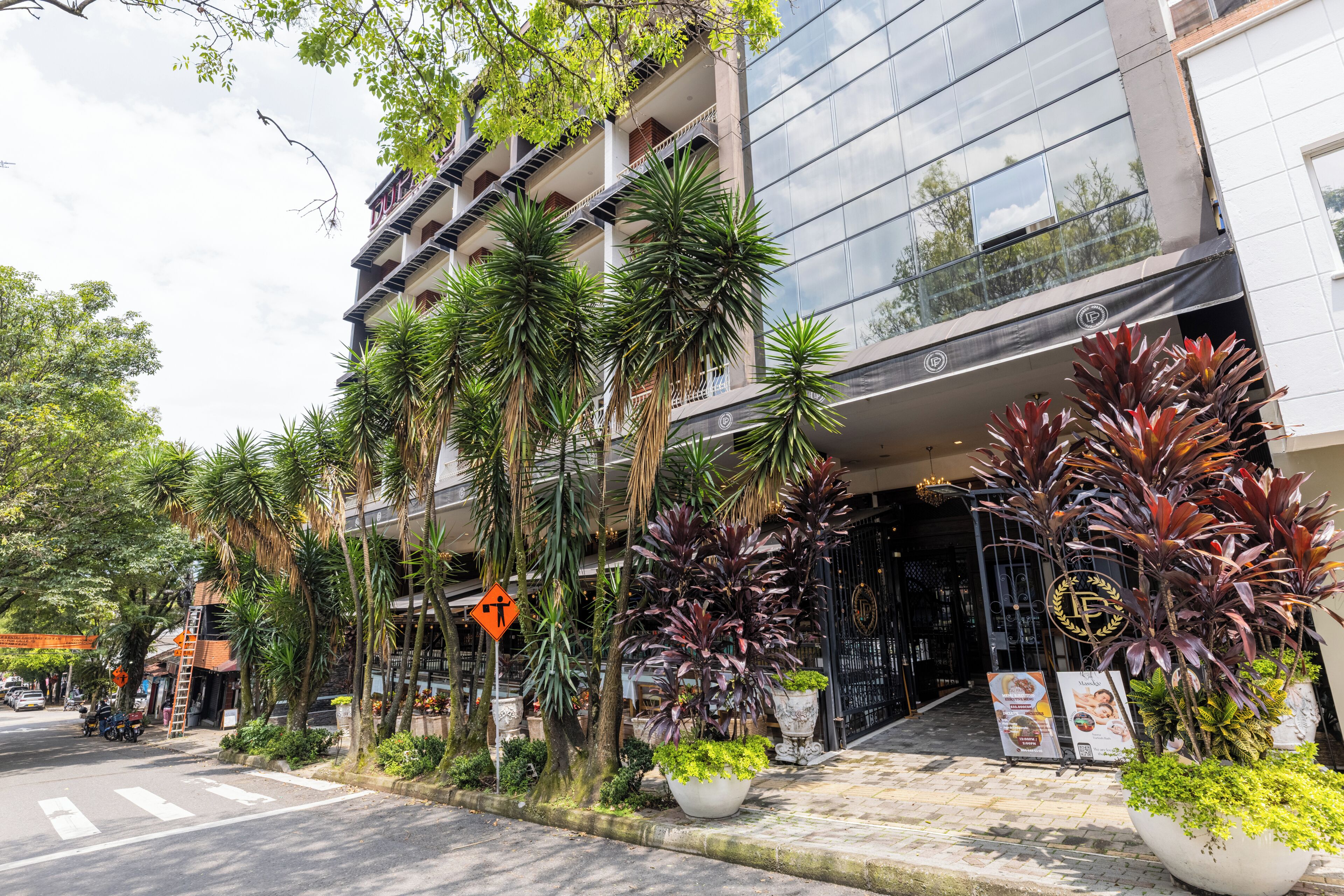 A modern building with a reflective glass facade, surrounded by lush tropical plants and a pedestrian crossing.