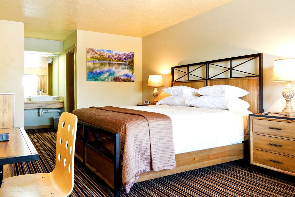 A hotel room with a bed, bedside table, lamp, chair, and a painting on the wall.