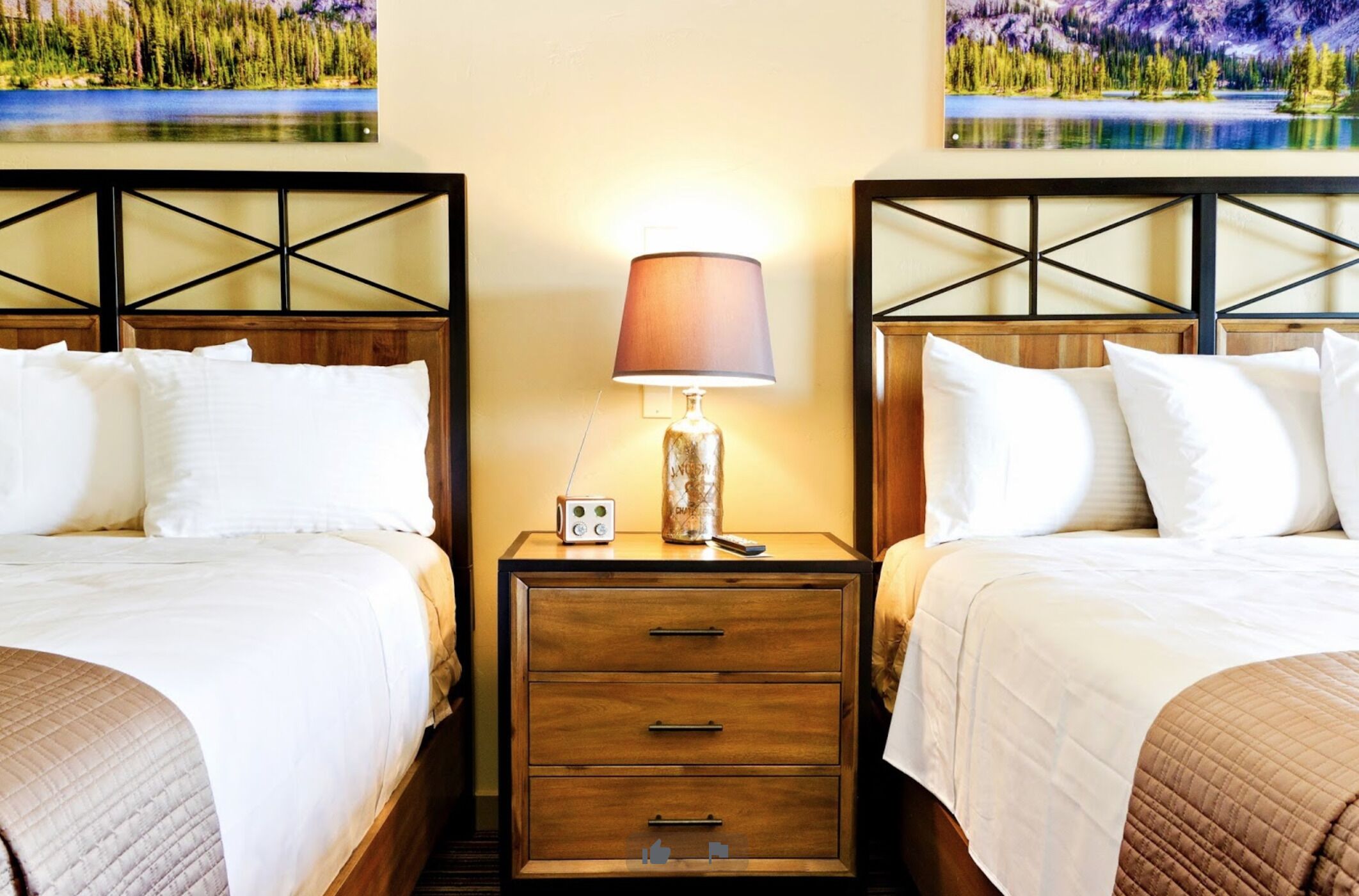 A hotel room with two beds, a nightstand with a lamp, and a painting of a lake.