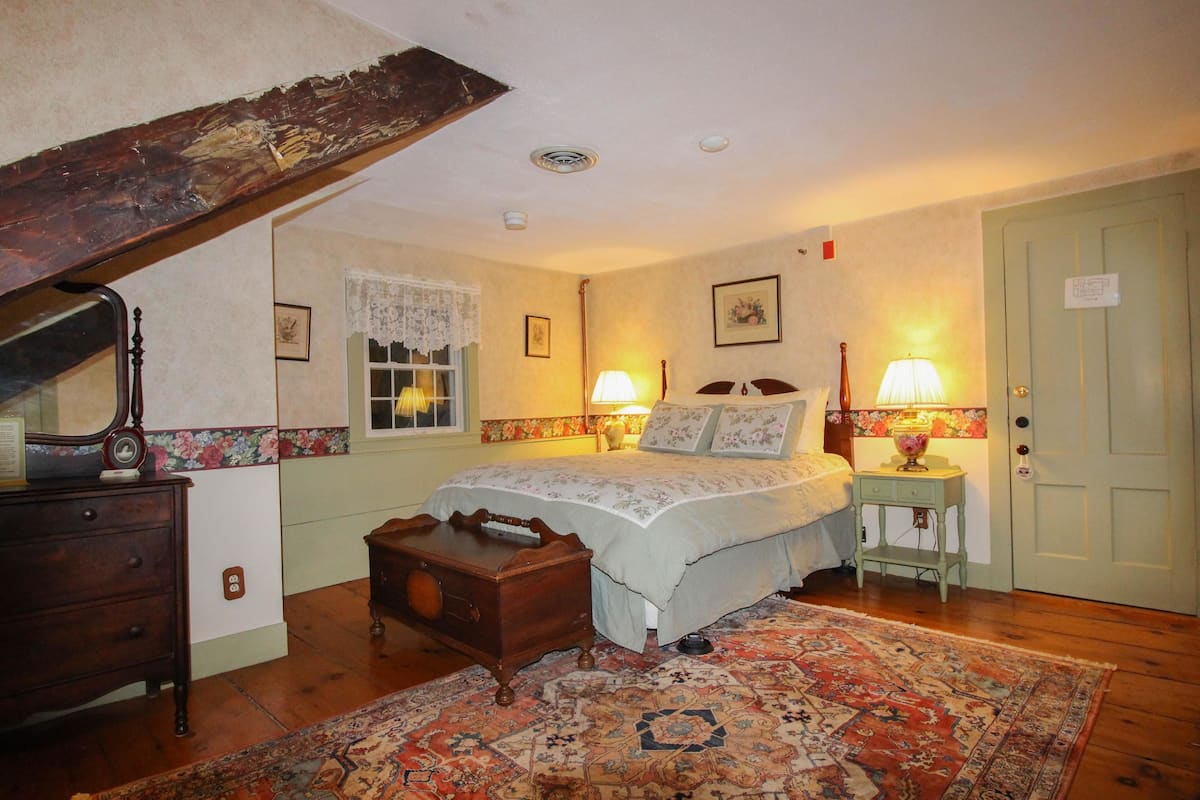 A bedroom with a bed, two bedside tables with lamps, a dresser, a wooden chest, a window with lace curtains, a door, and a patterned rug.