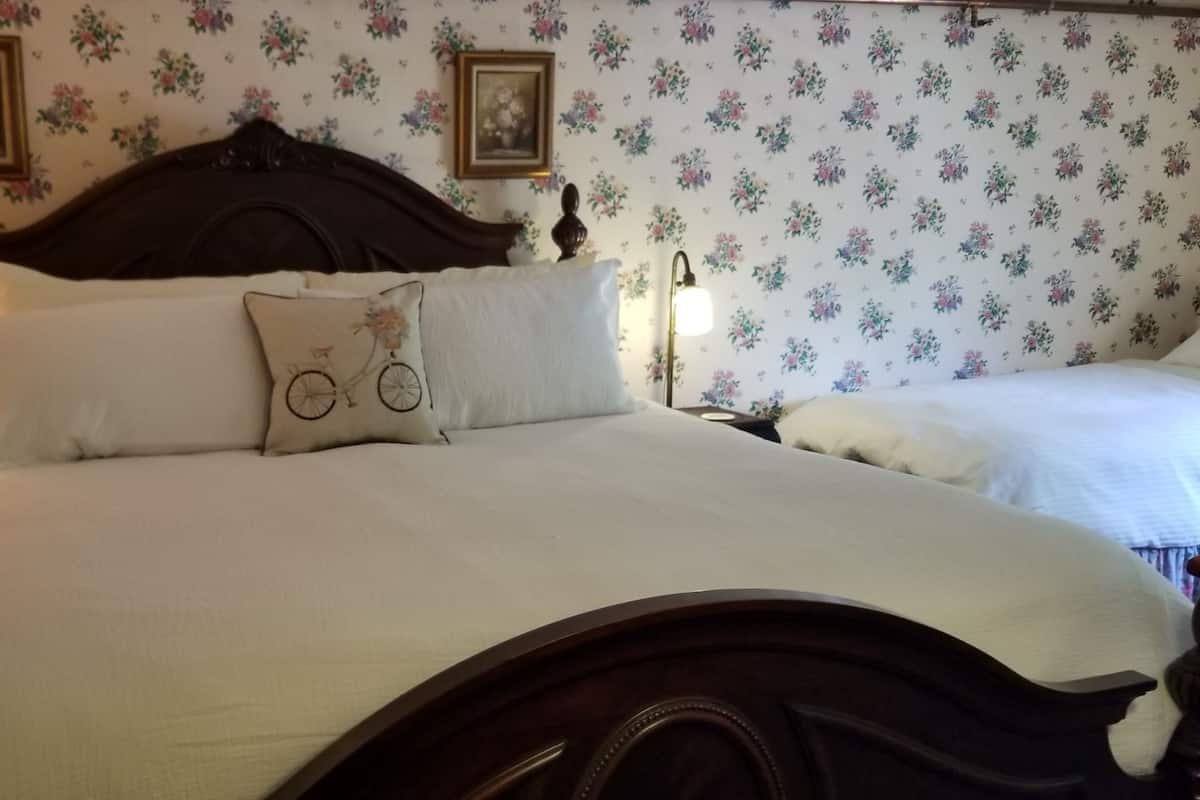 A bedroom with floral wallpaper, a wooden headboard, a bed with white bedding, two bedside lamps, and a nightstand with a lamp and a picture frame.