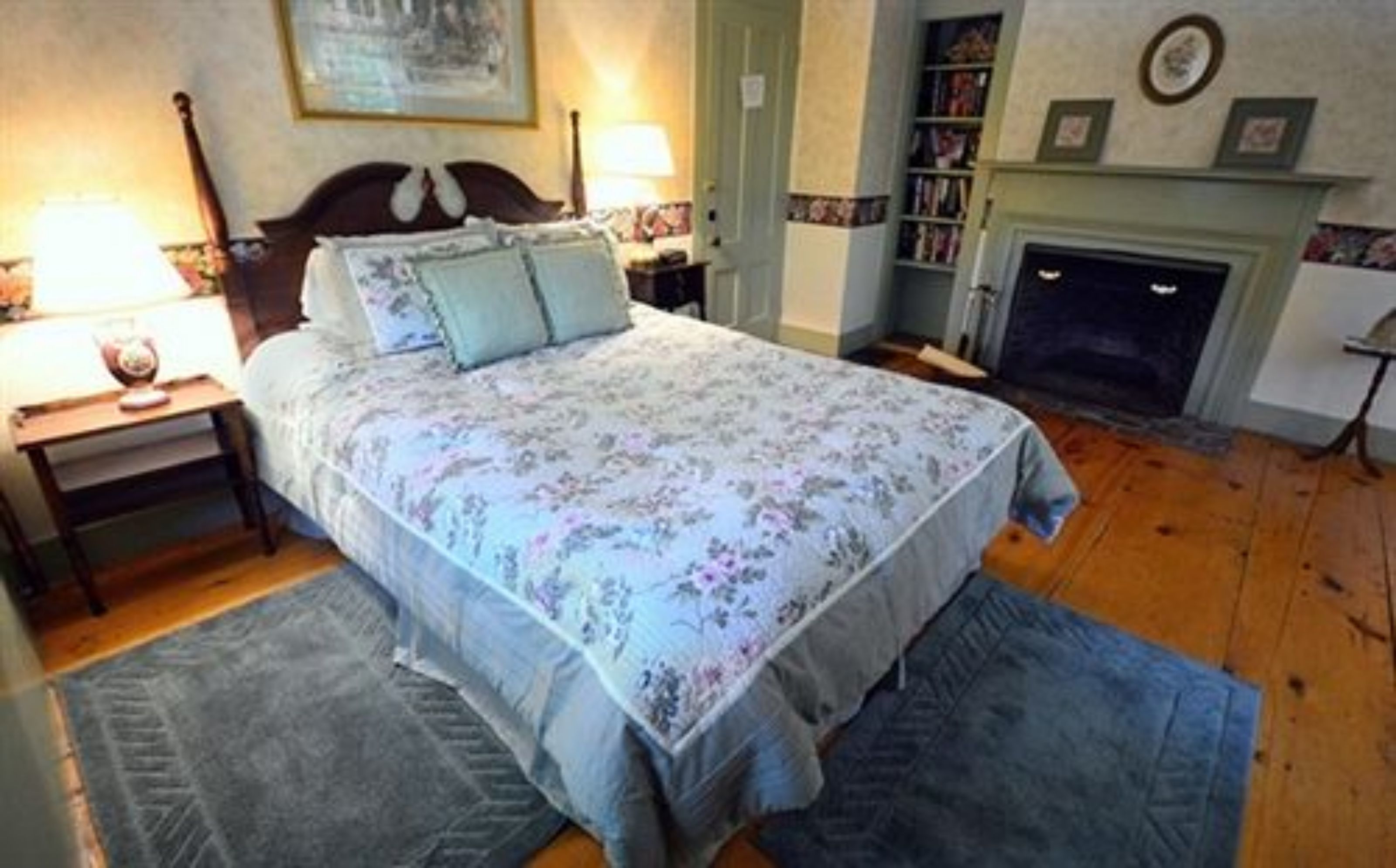 Quarto com colcha florida, piso de madeira, mesa de cabeceira com abajur, estante de livros e lareira.