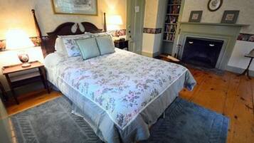 Quarto com colcha florida, piso de madeira, mesa de cabeceira com abajur, estante de livros e lareira.
