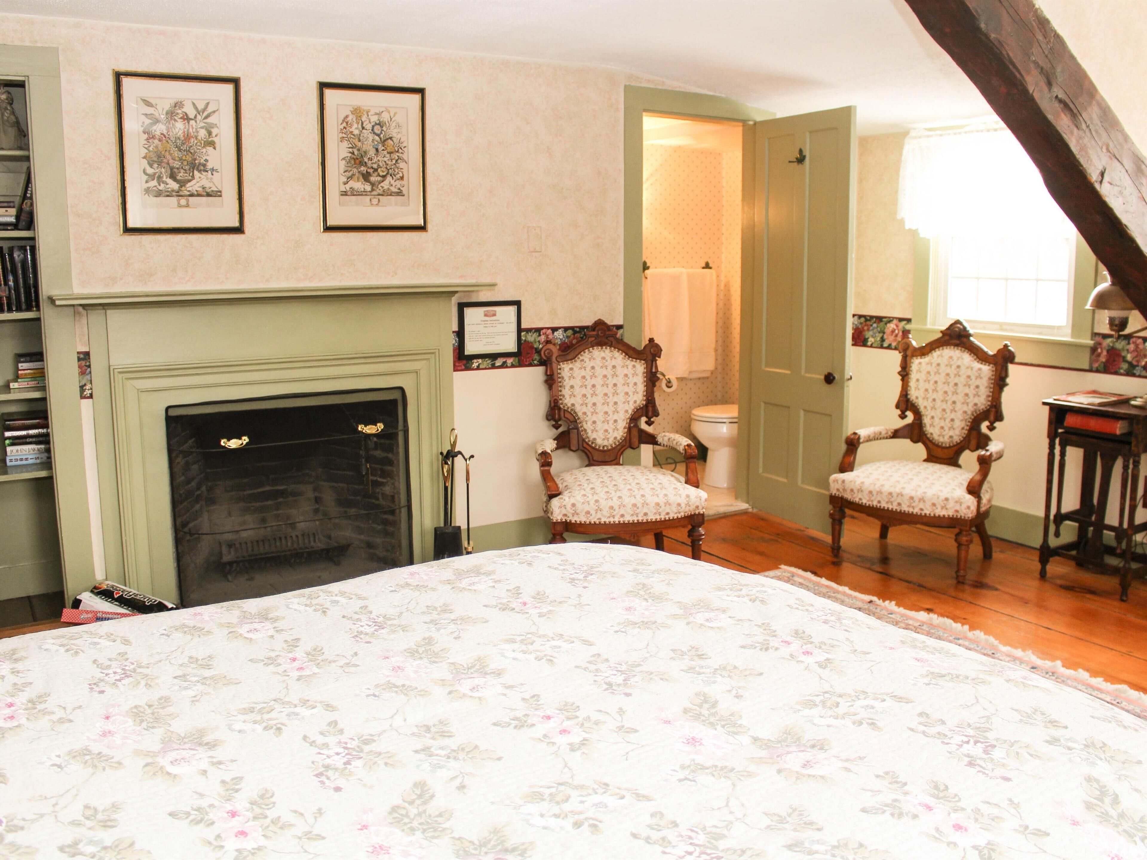 A room with a fireplace, two upholstered chairs, a bed with a floral bedspread, a bookshelf, and two framed floral paintings on the wall.