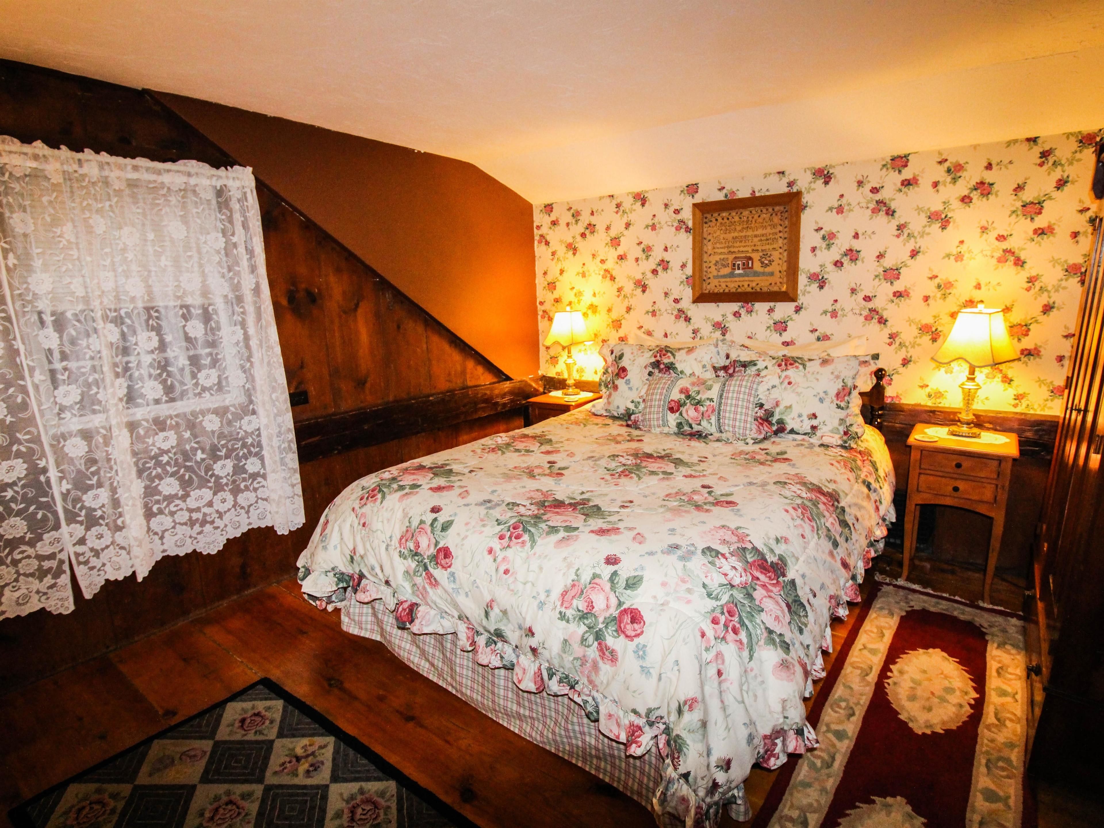 A bedroom with a wooden staircase, floral wallpaper, a bed with floral bedding, two bedside lamps, and a small bedside table.