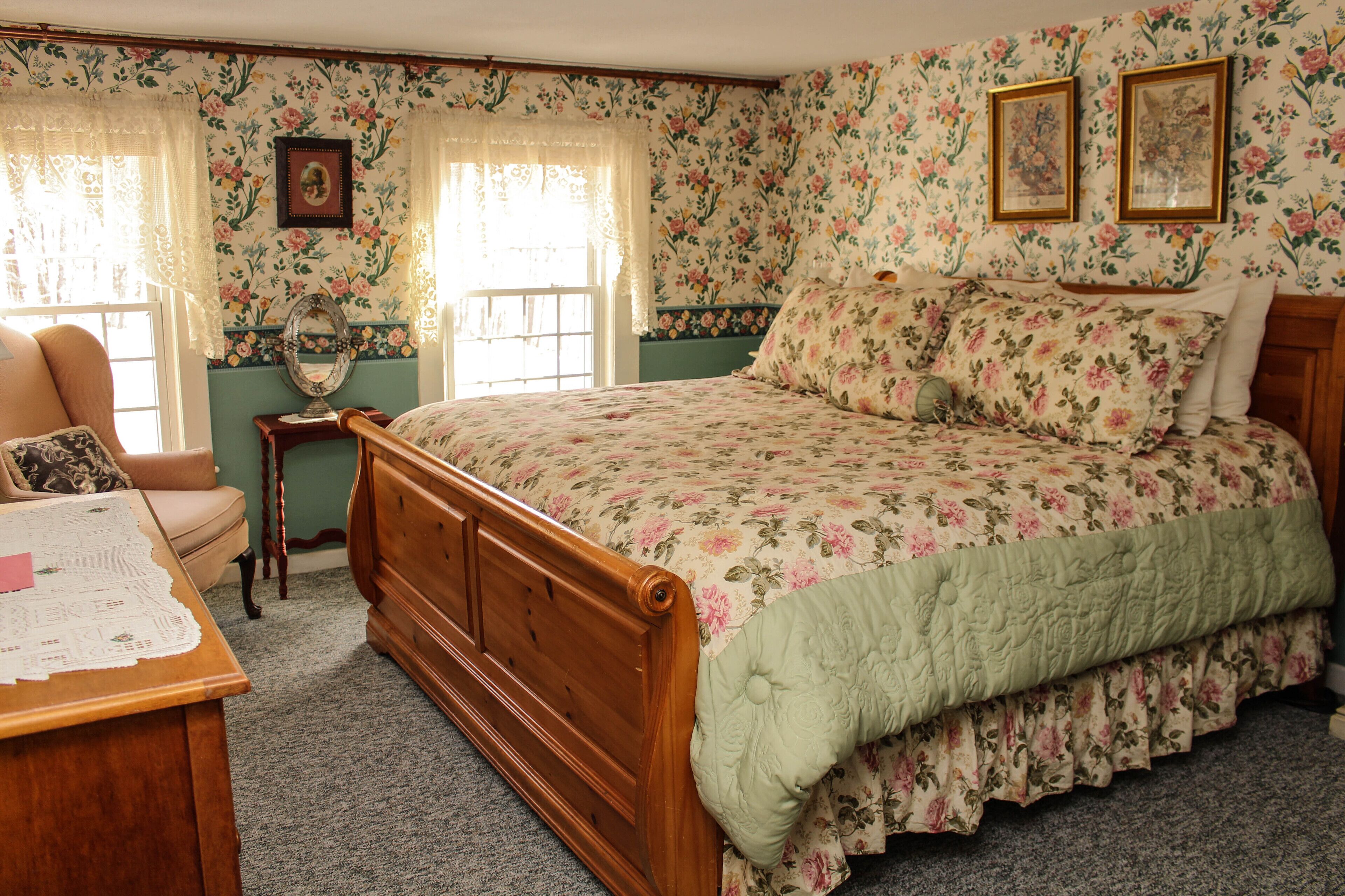 Un dormitorio con papel tapiz floral, una cama de madera, un escritorio con mantel de encaje y una silla.