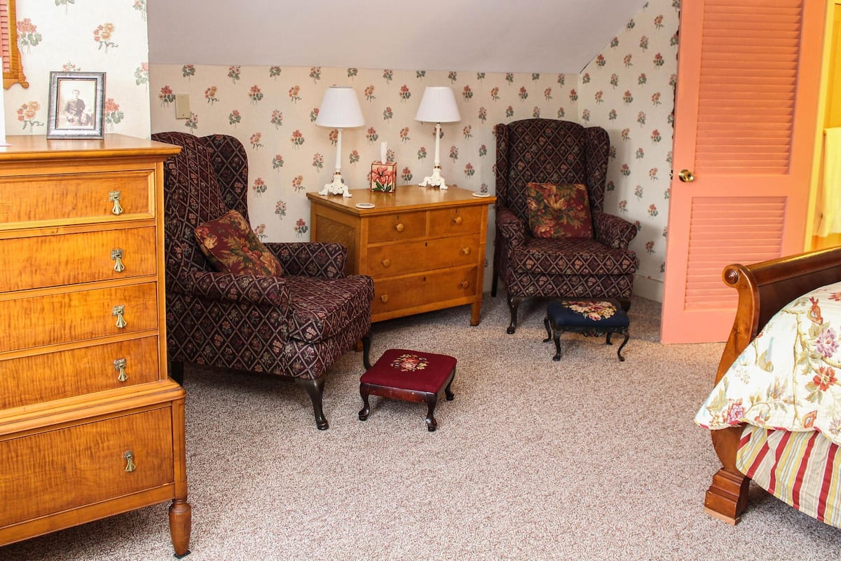 A room with a wooden dresser, two patterned armchairs, a wooden cabinet, and a small red ottoman.