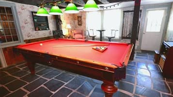 Uma mesa de bilhar com luzes verdes no teto, um relógio e uma porta em um cômodo com papel de parede estampado e piso de pedra.
