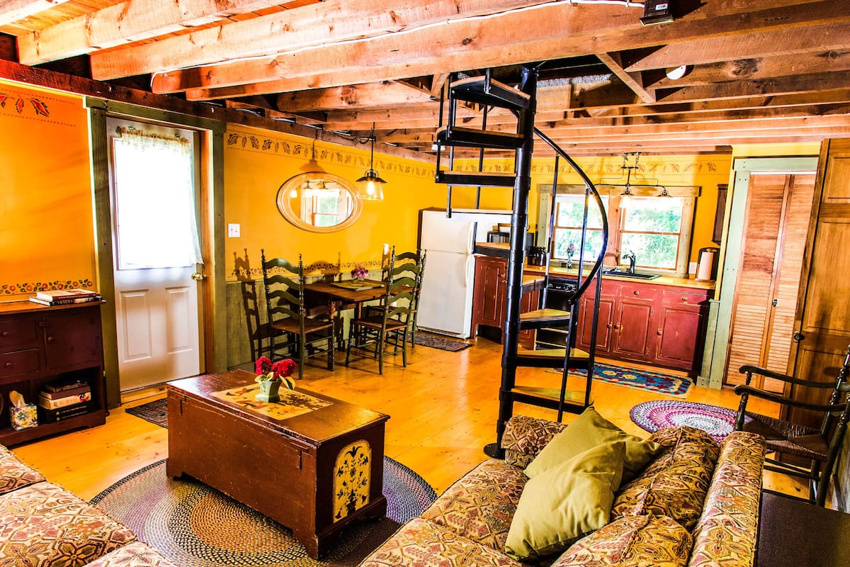 A cozy, rustic interior with a wooden ceiling, a dining area with a table and chairs, a kitchen with red cabinets, and a patterned rug on the floor.