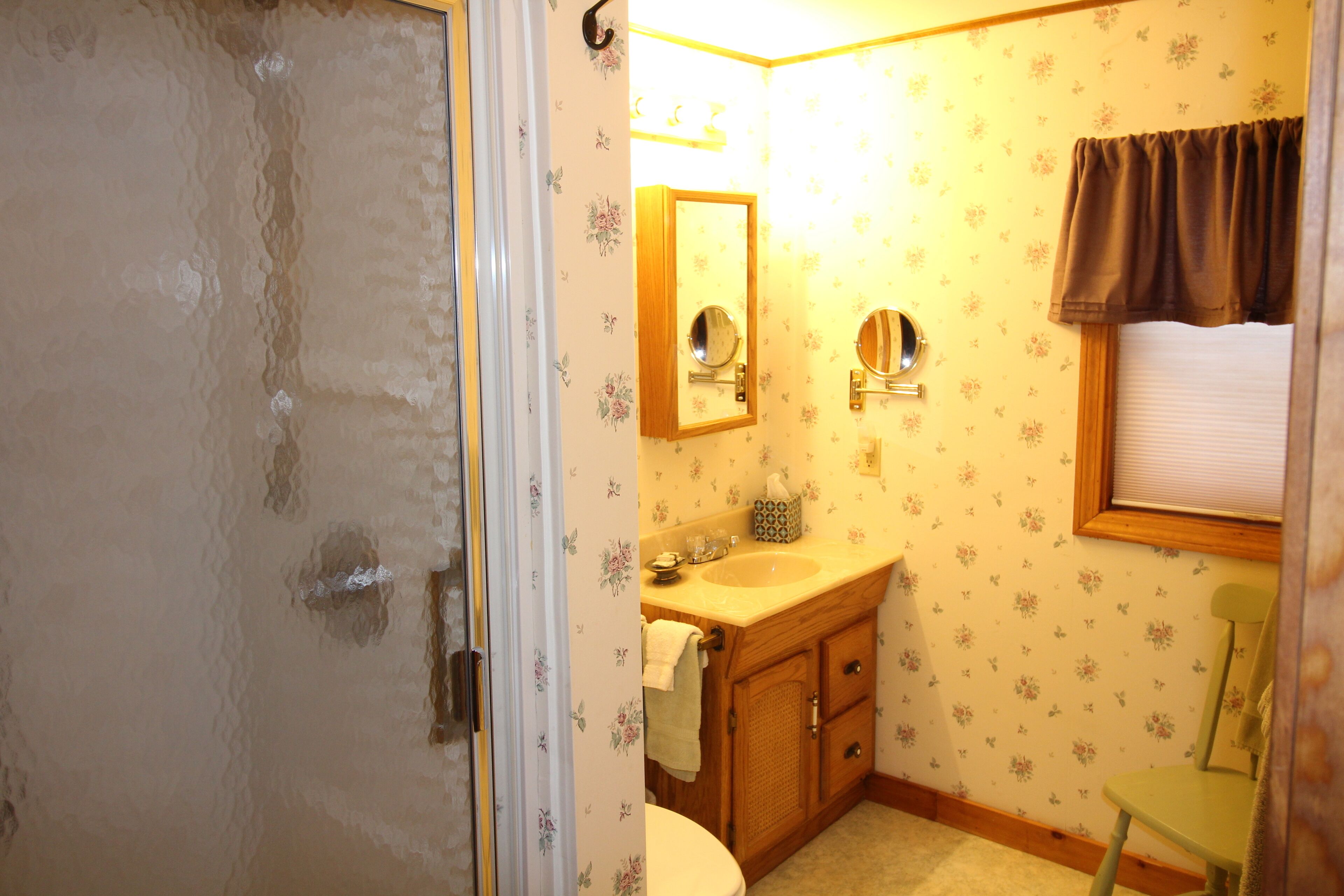 Ein Badezimmer mit einem Holzbadschrank, zwei Wandspiegeln, einer Toilette und einem Fenster mit brauner Vorhangstange.