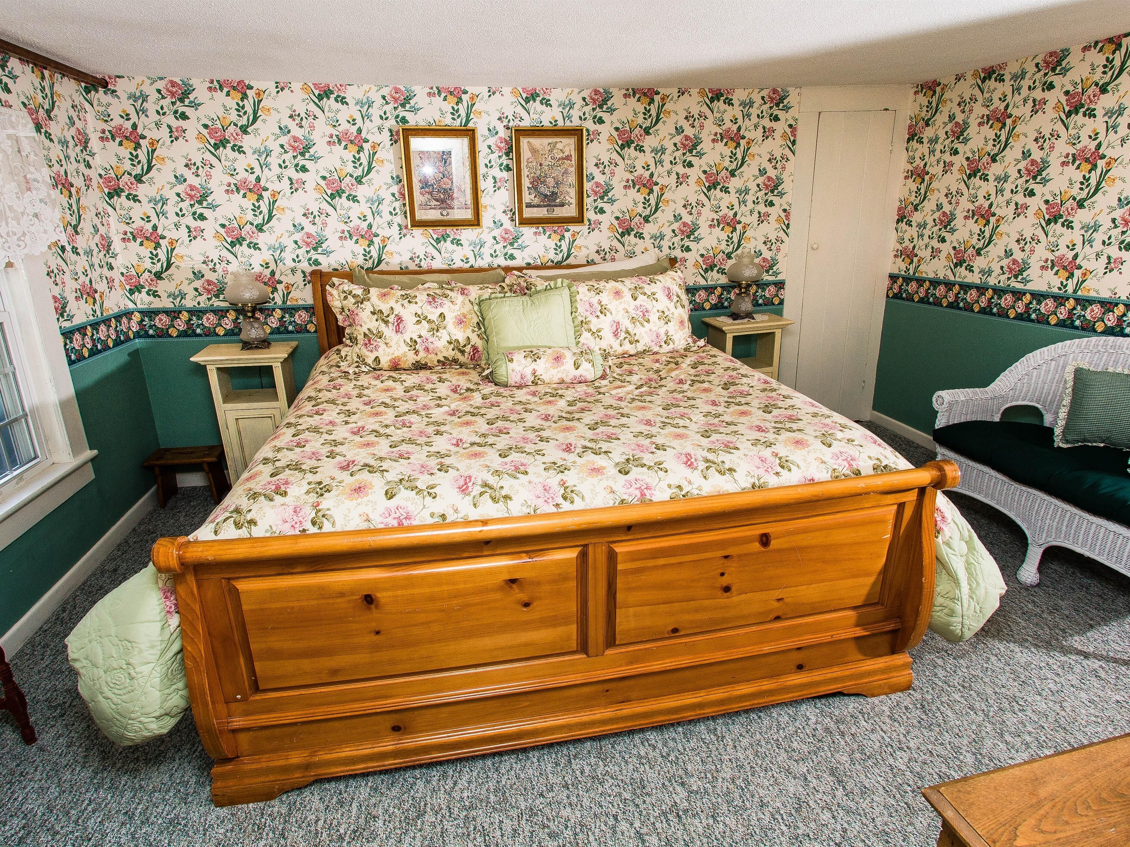 Une chambre à coucher avec une couvre-lit à motifs floraux, un cadre de lit en bois, une chaise en osier et deux tables de chevet avec des lampes.