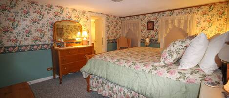 A bedroom with floral wallpaper, a bed with floral bedding, a dresser, a mirror, and a bedside table with a lamp.